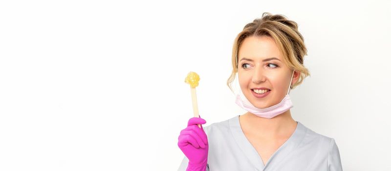 The master of sugar hair removal holds liquid yellow sugar paste, wax for depilation on a wooden stick on a white background