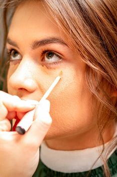 Beautiful young brunette woman receiving makeup with stick concealer on her face in a beauty salon