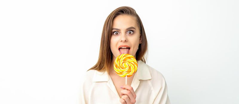 Beautiful young caucasian woman wearing a white shirt licking a lollipop on a white background