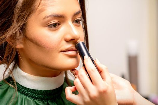 Beautiful young brunette woman receiving makeup with stick concealer on her face in a beauty salon