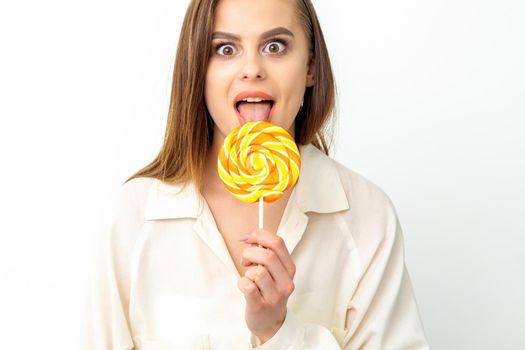 Beautiful young caucasian woman wearing a white shirt licking a lollipop on a white background