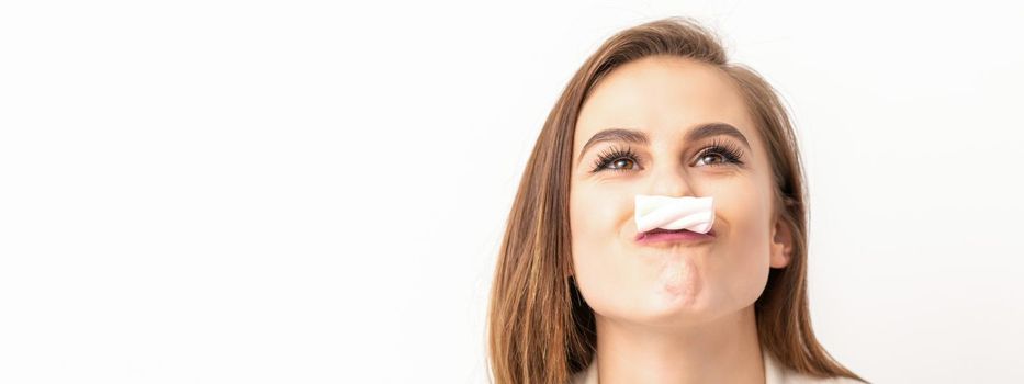 Funny young woman with marshmallow on her lips looking up and standing on white background with copy space