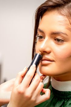 Beautiful young brunette woman receiving makeup with stick concealer on her face in a beauty salon