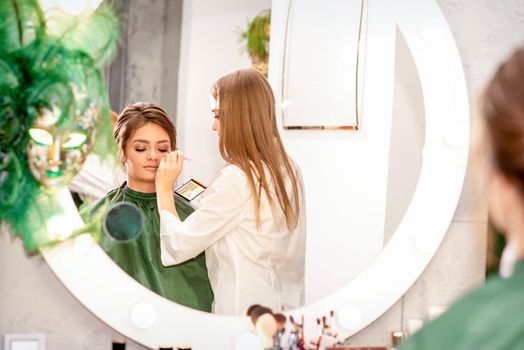 Professional makeup artist applies makeup to the eyes of the beautiful young woman in the mirror smiling in a beauty salon