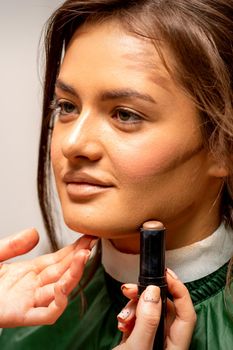 Beautiful young brunette woman receiving makeup with stick concealer on her face in a beauty salon