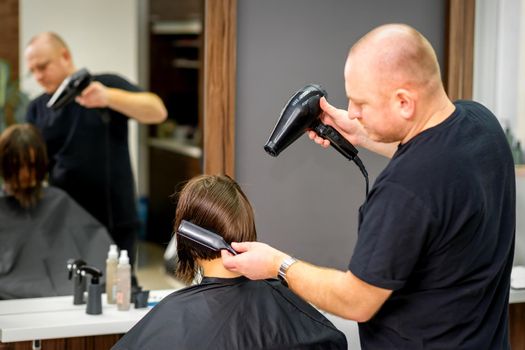 Male hairdresser drying short hair of young caucasian brunette woman with a black hairdryer and black round brush in a hairdresser salon