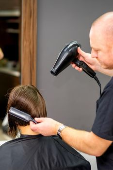 Male hairdresser drying short hair of young caucasian brunette woman with a black hairdryer and black round brush in a hairdresser salon