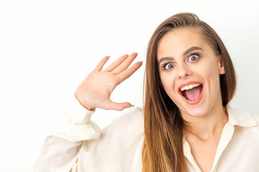 Portrait of young caucasian woman wearing white shirt raises hands and laughs positively with open mouth poses against a white background