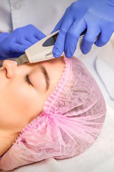Young caucasian woman receiving facial skin cleaning by ultrasonic cosmetology face equipment in a medical salon