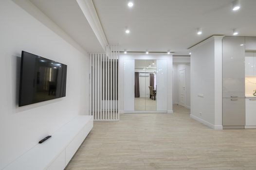 large white room connected to hall in a studio apartment, flat screen tv on the wall, part of white kitchen furniture at the very right side