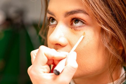 Beautiful young brunette woman receiving makeup with stick concealer on her face in a beauty salon