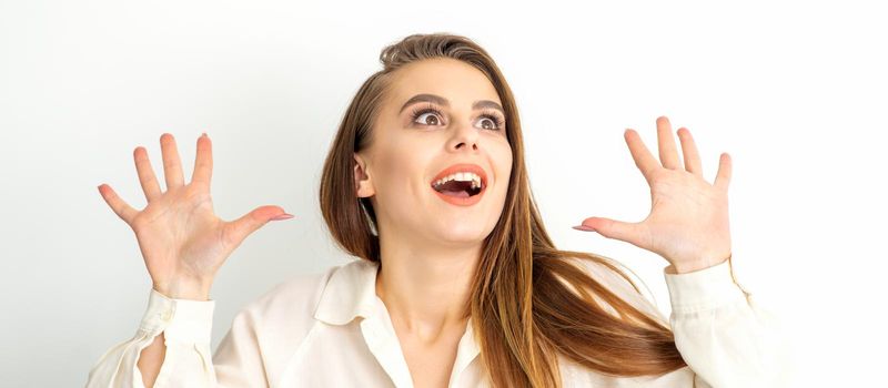 Portrait of young caucasian woman wearing white shirt raises hands and laughs positively with open mouth looking up against a white background