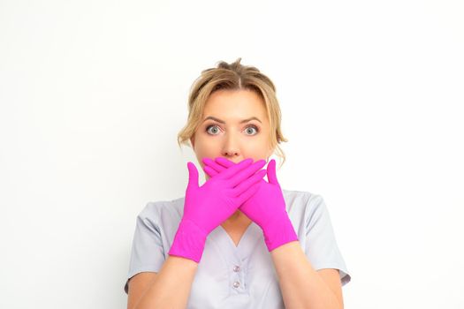 Portrait of a young female caucasian doctor or nurse is shocked covering her mouth with her pink gloved hands against a white background