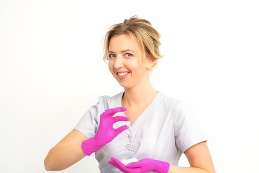 Portrait of a young smiling Caucasian beautician wearing pink gloves holding sugar cubes showing and looking at the camera against a white background