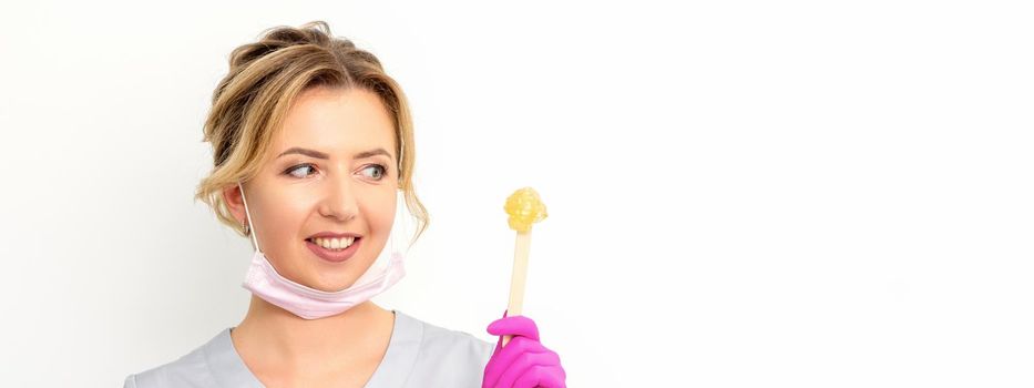 The master of sugar hair removal holds liquid yellow sugar paste, wax for depilation on a wooden stick on a white background