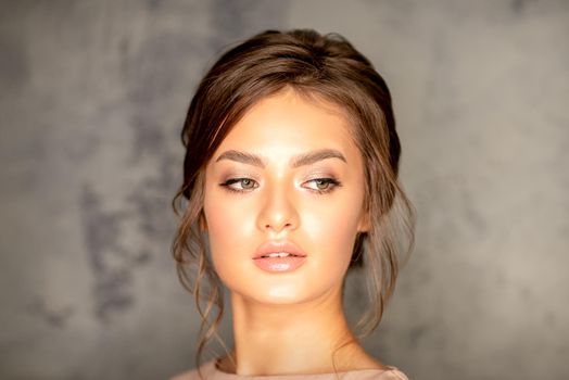 The fashionable young woman. Portrait of the beautiful female model with long hair and makeup. Beauty young caucasian woman with a brown curly hairstyle on the background of a gray wall