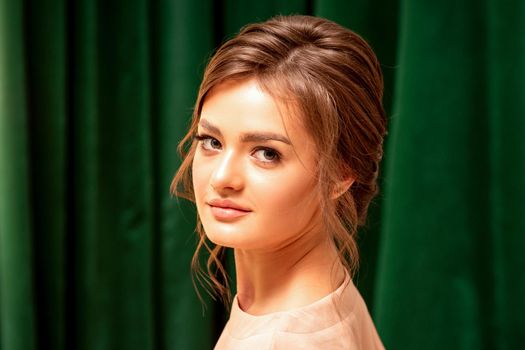 The fashionable young woman. Portrait of the beautiful female model with long hair and makeup with closed eyes. Beauty young woman with a brown curly hairstyle on the background of a green curtain