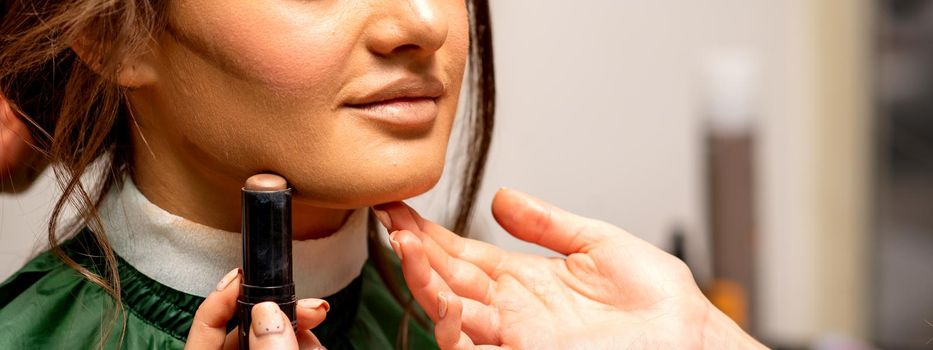 Beautiful young brunette woman receiving makeup with stick concealer on her face in a beauty salon