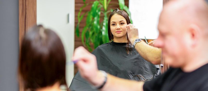 Reflection in the mirror of the young caucasian woman sitting and receiving haircut by male hairdresser at hairdresser salon