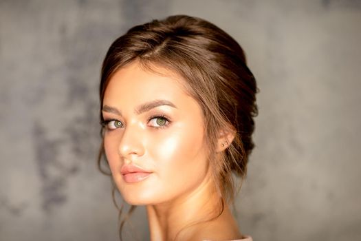 The fashionable young woman. Portrait of the beautiful female model with long hair and makeup. Beauty young caucasian woman with a brown curly hairstyle on the background of a gray wall