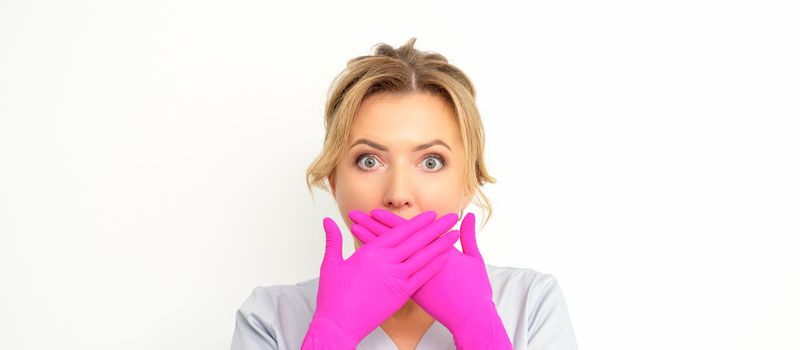 Portrait of a young female caucasian doctor or nurse is shocked covering her mouth with her pink gloved hands against a white background