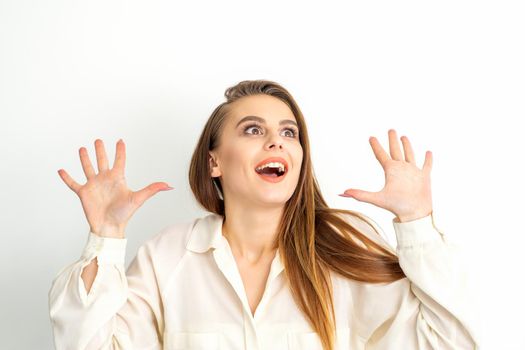 Portrait of young caucasian woman wearing white shirt raises hands and laughs positively with open mouth looking up against a white background