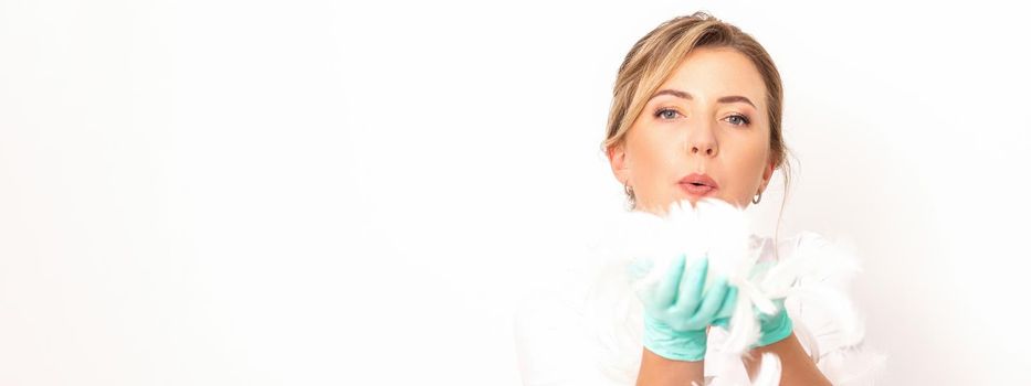 Young beautiful woman beautician in protective green gloves standing and blowing on feathers over white wall background