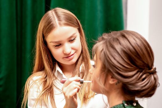 Makeup in the process. The makeup artist applies pink gloss lipstick on the lips of the beautiful face of the young caucasian woman in a beauty salon