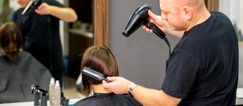 Male hairdresser drying short hair of young caucasian brunette woman with a black hairdryer and black round brush in a hairdresser salon