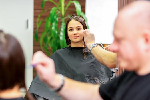 Reflection in the mirror of the young caucasian woman sitting and receiving haircut by male hairdresser at hairdresser salon