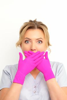 Portrait of a young female caucasian doctor or nurse is shocked covering her mouth with her pink gloved hands against a white background