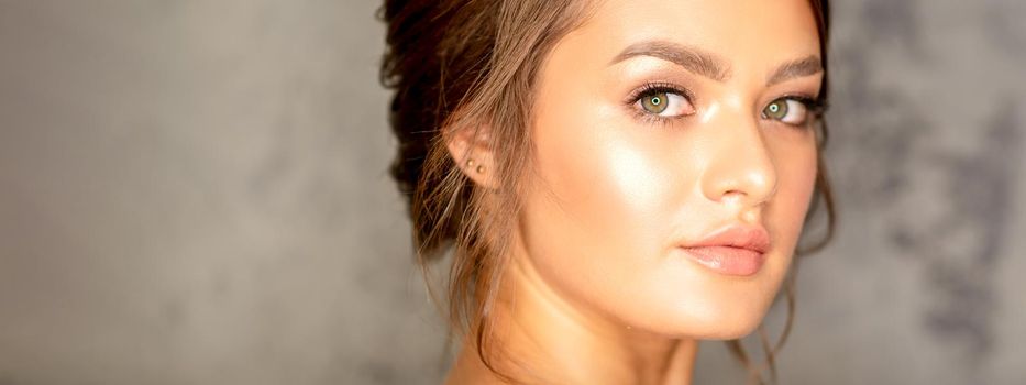 The fashionable young woman. Portrait of the beautiful female model with long hair and makeup. Beauty young caucasian woman with a brown curly hairstyle on the background of a gray wall