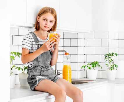 Preteen girl with orange juice bottle and glass sitting at kitchen table. Pretty child kid with vitamin fruit drink at home