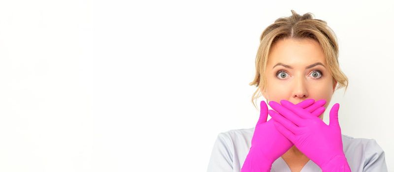 Portrait of a young female caucasian doctor or nurse is shocked covering her mouth with her pink gloved hands against a white background