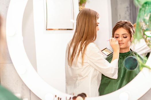 Professional makeup artist applies makeup to the eyes of the beautiful young woman in the mirror smiling in a beauty salon