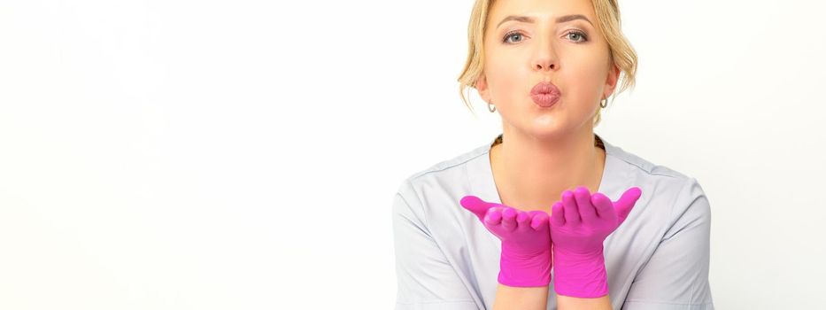 Young Caucasian doctor woman wearing pink gloves folding lips and holding palms to send air kiss isolated white background