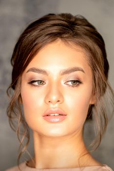 The fashionable young woman. Portrait of the beautiful female model with long hair and makeup. Beauty young caucasian woman with a brown curly hairstyle on the background of a gray wall