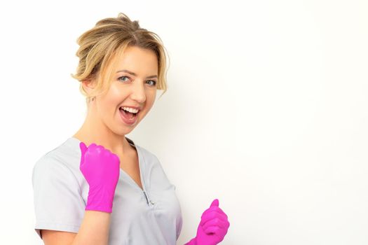 Happy caucasian woman doctor wearing pink gloves celebrates and raising fists on white background