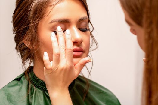 The hand of a makeup artist applies eye shadow on the eyelid of a young Caucasian woman with fingers in a beauty salon