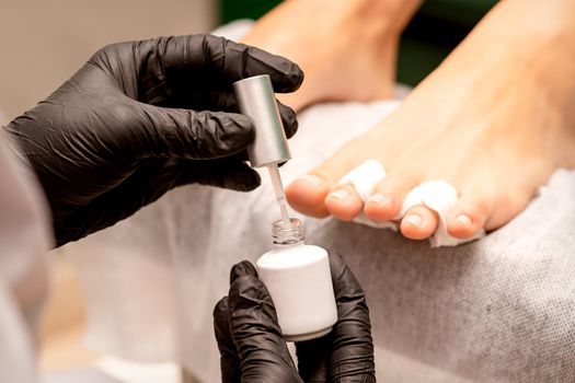 White nail polish in the hands of a manicurist while painting nails on a female feet, closeup