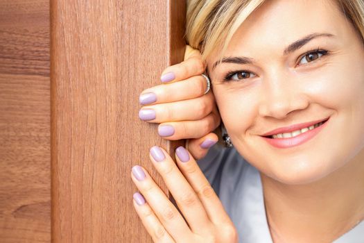 Close up portrait of the young smiling caucasian woman looks out from behind the door looking at the camera