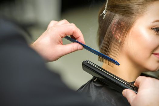 Hairstylist is straightening short hair of young brunette woman with a flat iron in a hairdresser salon, close up