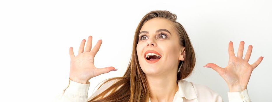 Portrait of young caucasian woman wearing white shirt raises hands and laughs positively with open mouth looking up against a white background