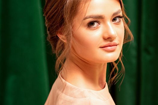 The fashionable young woman. Portrait of the beautiful female model with long hair and makeup with closed eyes. Beauty young woman with a brown curly hairstyle on the background of a green curtain