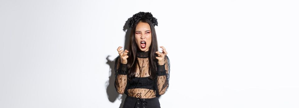 Image of evil witch hate something, looking angry and furious at upper left corner, cursing or swearing while clenching fists mad, standing in halloween costume over white background.