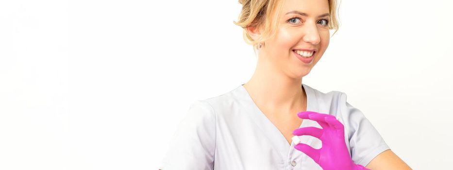 Portrait of a young smiling Caucasian beautician wearing pink gloves holding sugar cubes showing and looking at the camera against a white background