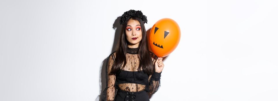 Image of silly beautiful asian woman in witch costume, holding orange balloon with scary face, celebrating halloween.