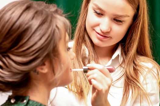 Makeup in the process. The makeup artist applies pink gloss lipstick on the lips of the beautiful face of the young caucasian woman in a beauty salon