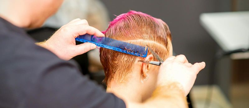 Haircut of dyed short pink wet hair of young caucasian woman by a male hairdresser in a barbershop