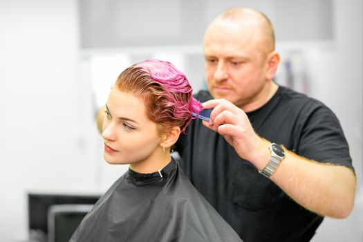 Haircut of dyed short pink wet hair of young caucasian woman by a male hairdresser in a barbershop